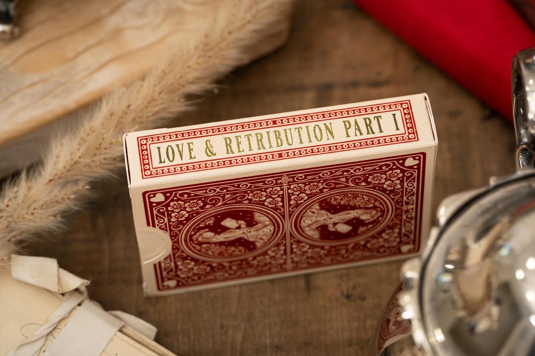 Playing card box titled 'Love & Retribution Part I' on a wooden surface with a red object and metal object in the background.
