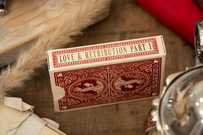 Playing card box titled 'Love & Retribution Part I' on a wooden surface with a red object and metal object in the background.