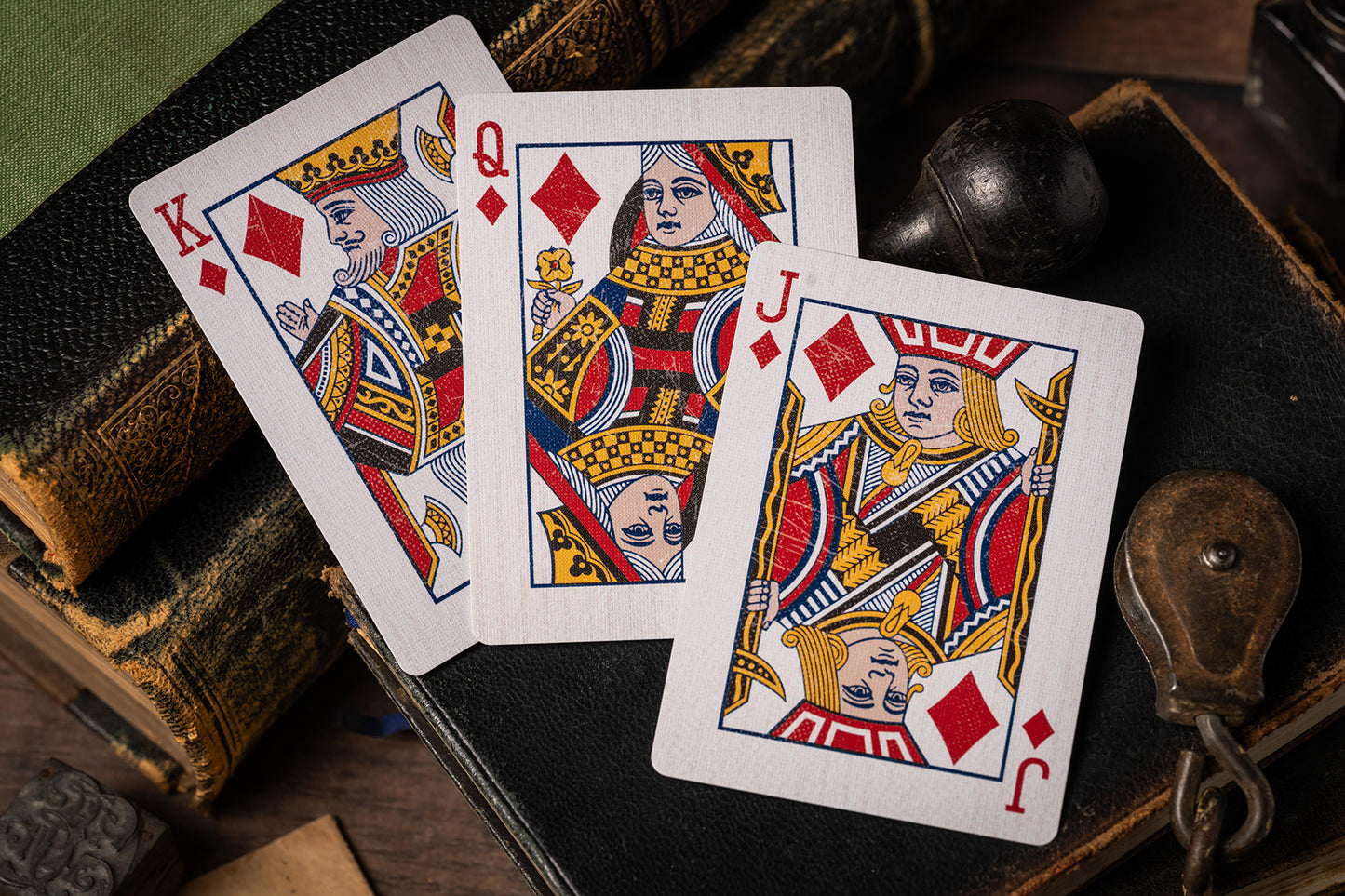 Three playing cards (King, Queen, and Jack of Diamonds) on a wooden surface with vintage books and leather-bound box.
