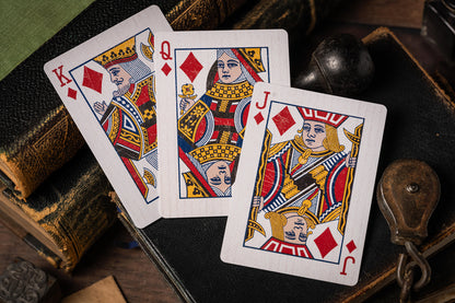 Three playing cards (King, Queen, and Jack of Diamonds) on a wooden surface with vintage books and leather-bound box.