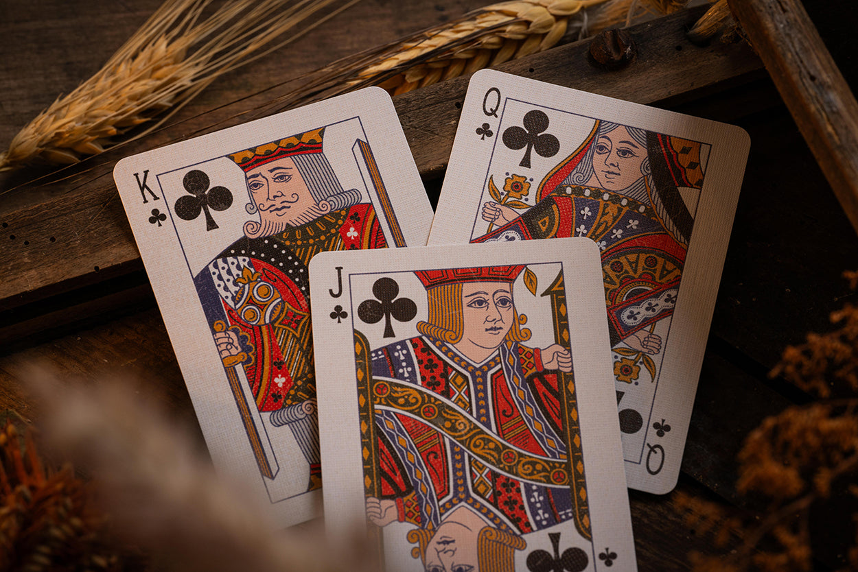 Four playing cards (King, Queen, Jack, of Clubs) on a wooden surface with wheat stalks.