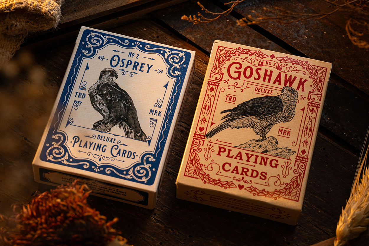 Two playing card decks, one blue and one red, with bird illustrations on wooden surface. hand illustrated in the UK